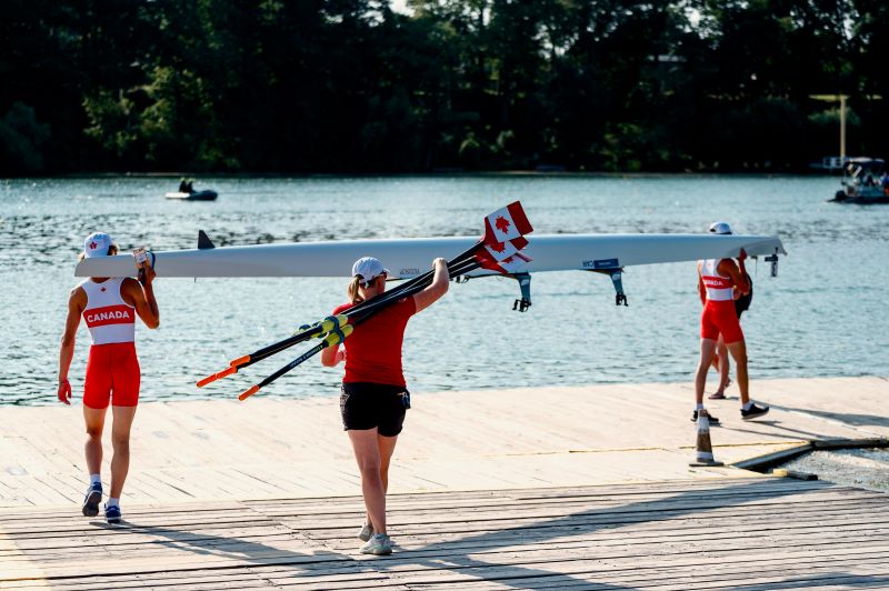 Two rowers launch a HUDSON single from a dock. A parent is carrying the oars with the Canadian maple leaf design.